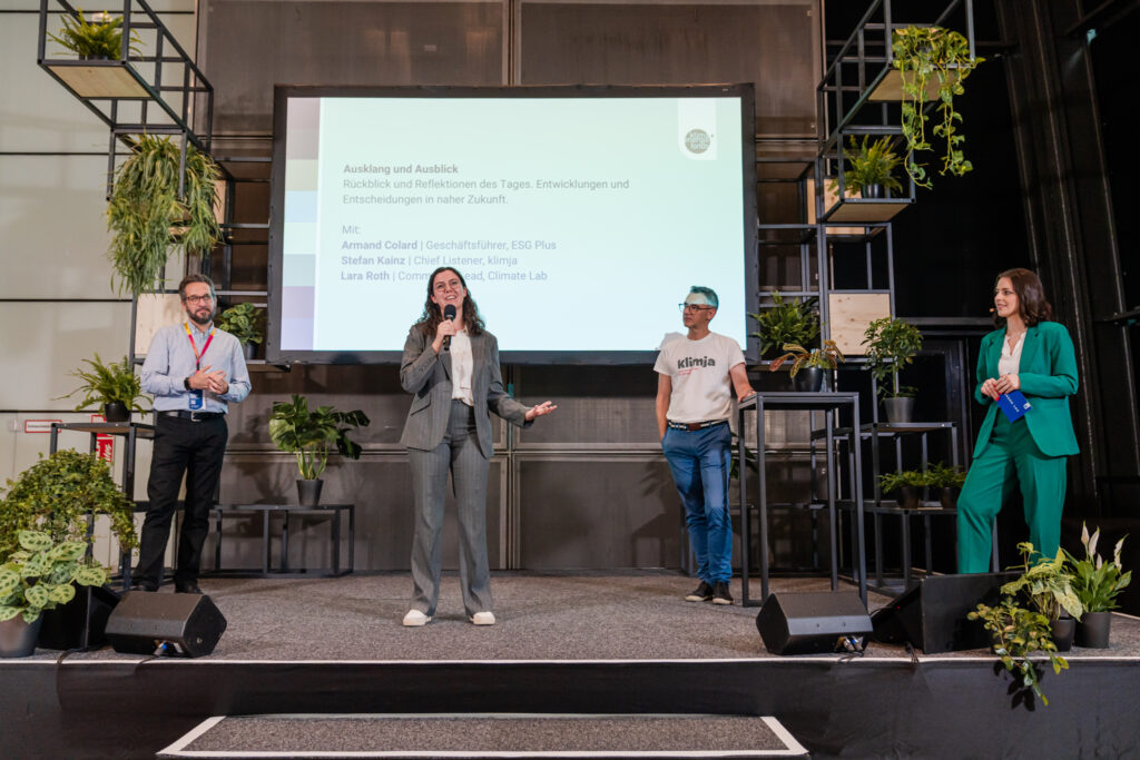 Das Bild zeigt eine Podiumsdiskussion auf einer modern gestalteten Bühne mit grünen Pflanzen und schwarzen Metallregalen. Eine Frau in einem grauen Anzug spricht ins Mikrofon, während drei weitere Personen auf der Bühne stehen und zuhören. Einer der Männer trägt ein weißes T-Shirt mit der Aufschrift „klimja“, während die Frau rechts in einem grünen Hosenanzug ein blaues Heft hält. Die große Leinwand im Hintergrund zeigt den Titel „Ausklang und Ausblick“ sowie die Namen der Diskussionsteilnehmer, darunter Armand Colard, Stefan Kainz und Lara Roth. Die Szene vermittelt eine professionelle, aber entspannte Atmosphäre in einem nachhaltigen Event-Setting.