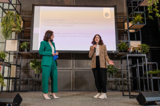 Das Bild zeigt zwei Frauen auf einer Bühne, die an einer Präsentation teilnehmen. Die Frau auf der linken Seite trägt einen eleganten grünen Hosenanzug mit silbernen High Heels und hält ein blaues Heft in der Hand. Die Frau auf der rechten Seite spricht ins Mikrofon und trägt einen karierten Blazer mit schwarzer Hose und weißen Sneakers. Im Hintergrund ist eine große Leinwand mit Text zu sehen, auf der Begriffe wie „Rückblick und Ausblick“ sowie das „Bundesministerium für Klimaschutz“ zu erkennen sind. Die Bühne ist mit modernen schwarzen Metallregalen und zahlreichen grünen Pflanzen dekoriert, was eine nachhaltige und stilvolle Atmosphäre schafft.