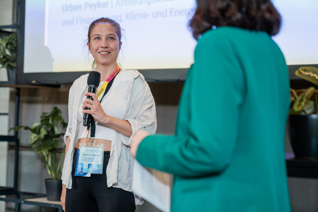 Das Bild zeigt eine Frau auf einer Bühne, die ein Mikrofon hält und lächelnd spricht. Sie trägt ein Namensschild an einem bunten Lanyard und ist in ein helles, lässiges Outfit gekleidet. Im Vordergrund ist eine andere Person in einem grünen Blazer zu sehen, die ein Blatt Papier hält und offenbar an der Konversation beteiligt ist. Der Hintergrund besteht aus einer Leinwand mit einer Präsentation und einigen Pflanzen, die eine nachhaltige Atmosphäre vermitteln.
