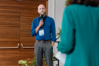 Das Bild zeigt einen Mann in einem blauen Hemd, der auf einer Bühne steht und ein Mikrofon hält. Er trägt ein Namensschild an einem schwarzen Lanyard und scheint eine Rede oder Präsentation zu halten. Im Vordergrund ist eine Person in einem grünen Blazer zu sehen, die ihm zuhört und ein Blatt Papier in der Hand hält. Der Hintergrund besteht aus einer Holzvertäfelung und mehreren Pflanzen, was eine nachhaltige und natürliche Atmosphäre schafft.