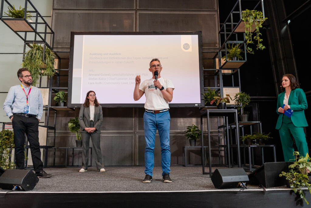 Das Bild zeigt eine Podiumsdiskussion oder Abschlussrede auf einer nachhaltigkeitsorientierten Veranstaltung. Ein Mann in einem weißen „klima“-T-Shirt spricht ins Mikrofon und gestikuliert mit der Hand. Hinter ihm ist eine große Präsentationsfolie mit Text zu sehen, die unter anderem Namen von Diskussionsteilnehmenden nennt: Armand Colard, Stefan Kainz und Lara Roth. Links auf der Bühne stehen zwei weitere Personen in Business-Outfits, während rechts eine Frau in einem grünen Hosenanzug mit einem blauen Moderationskärtchen steht. Die Bühne ist mit vielen Pflanzen dekoriert, und das moderne Design unterstreicht das nachhaltige Thema der Veranstaltung.