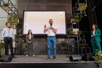 Das Bild zeigt eine Podiumsdiskussion oder Abschlussrede auf einer nachhaltigkeitsorientierten Veranstaltung. Ein Mann in einem weißen „klima“-T-Shirt spricht ins Mikrofon und gestikuliert mit der Hand. Hinter ihm ist eine große Präsentationsfolie mit Text zu sehen, die unter anderem Namen von Diskussionsteilnehmenden nennt: Armand Colard, Stefan Kainz und Lara Roth. Links auf der Bühne stehen zwei weitere Personen in Business-Outfits, während rechts eine Frau in einem grünen Hosenanzug mit einem blauen Moderationskärtchen steht. Die Bühne ist mit vielen Pflanzen dekoriert, und das moderne Design unterstreicht das nachhaltige Thema der Veranstaltung.