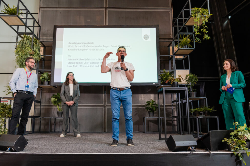 Das Bild zeigt eine Podiumsdiskussion oder Abschlussrede auf einer nachhaltigkeitsorientierten Veranstaltung. Ein Mann in einem weißen „klima“-T-Shirt spricht ins Mikrofon und gestikuliert mit der Hand. Hinter ihm ist eine große Präsentationsfolie mit Text zu sehen, die unter anderem Namen von Diskussionsteilnehmenden nennt: Armand Colard, Stefan Kainz und Lara Roth. Links auf der Bühne stehen zwei weitere Personen in Business-Outfits, während rechts eine Frau in einem grünen Hosenanzug mit einem blauen Moderationskärtchen steht. Die Bühne ist mit vielen Pflanzen dekoriert, und das moderne Design unterstreicht das nachhaltige Thema der Veranstaltung.