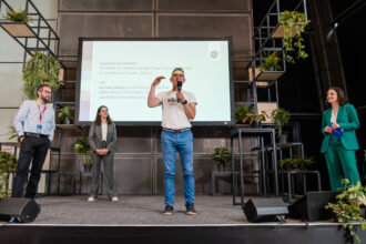 Das Bild zeigt eine Podiumsdiskussion oder Abschlussrede auf einer nachhaltigkeitsorientierten Veranstaltung. Ein Mann in einem weißen „klima“-T-Shirt spricht ins Mikrofon und gestikuliert mit der Hand. Hinter ihm ist eine große Präsentationsfolie mit Text zu sehen, die unter anderem Namen von Diskussionsteilnehmenden nennt: Armand Colard, Stefan Kainz und Lara Roth. Links auf der Bühne stehen zwei weitere Personen in Business-Outfits, während rechts eine Frau in einem grünen Hosenanzug mit einem blauen Moderationskärtchen steht. Die Bühne ist mit vielen Pflanzen dekoriert, und das moderne Design unterstreicht das nachhaltige Thema der Veranstaltung.