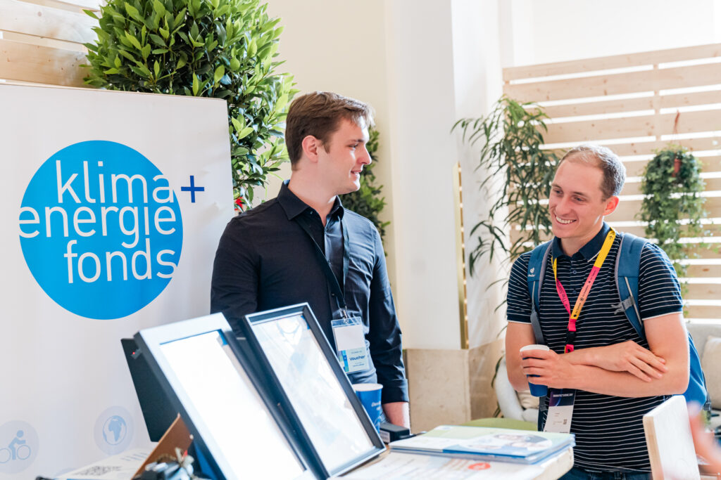 Das Bild zeigt zwei Männer an einem Infostand des Klima- und Energiefonds auf einer Veranstaltung. Der Mann links trägt ein schwarzes Hemd und hat ein Namensschild um den Hals, während der Mann rechts ein gestreiftes Poloshirt, einen Rucksack und eine bunte Akkreditierung trägt. Er hält eine Kaffeetasse in der Hand und lächelt während des Gesprächs. Im Vordergrund sind digitale Infotafeln und Broschüren sichtbar. Die Szene wirkt professionell, aber entspannt, mit einer nachhaltigen und umweltfreundlichen Atmosphäre durch Pflanzen und Holzwände im Hintergrund.