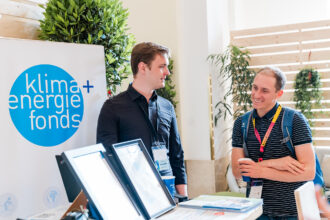 Das Bild zeigt zwei Männer an einem Infostand des Klima- und Energiefonds auf einer Veranstaltung. Der Mann links trägt ein schwarzes Hemd und hat ein Namensschild um den Hals, während der Mann rechts ein gestreiftes Poloshirt, einen Rucksack und eine bunte Akkreditierung trägt. Er hält eine Kaffeetasse in der Hand und lächelt während des Gesprächs. Im Vordergrund sind digitale Infotafeln und Broschüren sichtbar. Die Szene wirkt professionell, aber entspannt, mit einer nachhaltigen und umweltfreundlichen Atmosphäre durch Pflanzen und Holzwände im Hintergrund.
