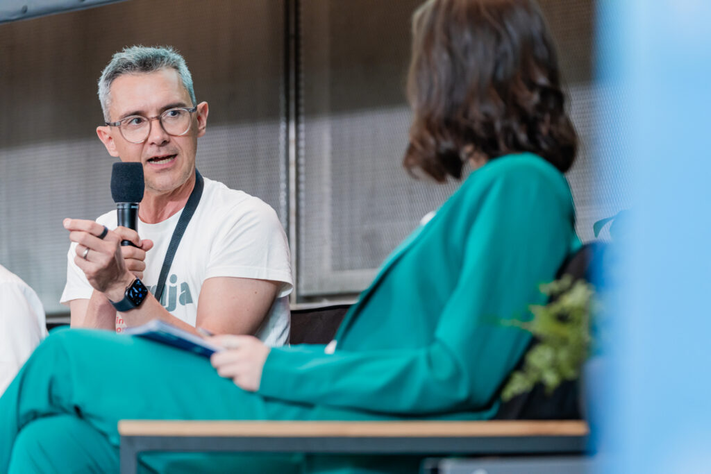 Das Bild zeigt eine Panel-Diskussion, bei der ein Mann mit Brille, grauen Haaren und einem weißen T-Shirt mit grünem Schriftzug in ein Mikrofon spricht. Er trägt eine Smartwatch und macht eine erklärende Handbewegung. Ihm gegenüber sitzt eine Frau in einem grünen Anzug, die ihm zuhört und Notizen hält. Die Szene wirkt professionell und findet vor einem metallischen Hintergrund statt, mit einem modernen Bühnen-Setup.