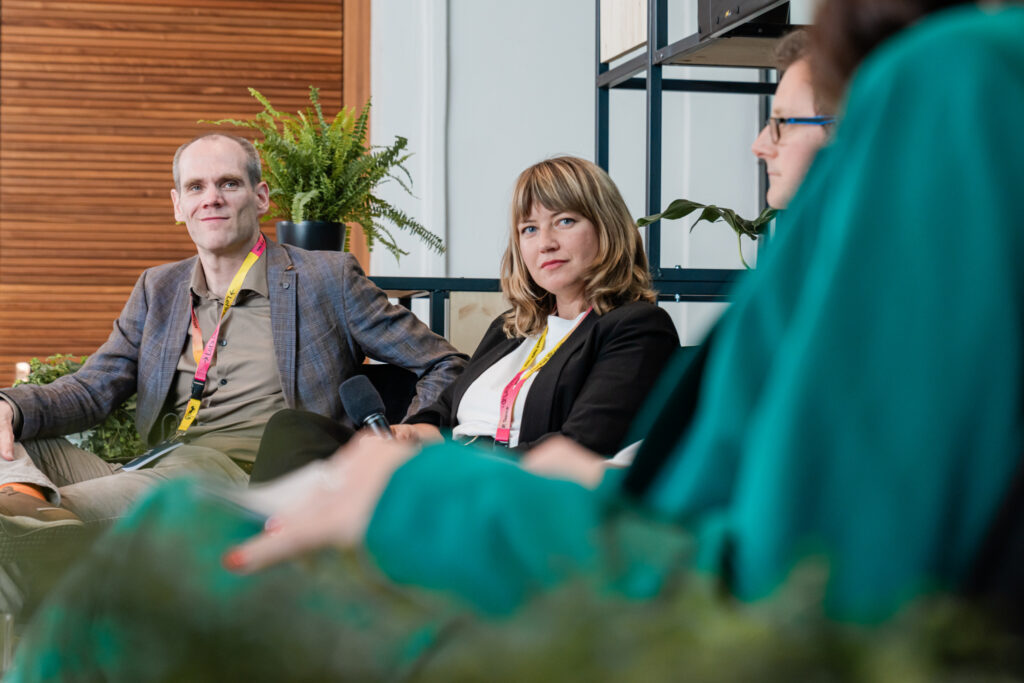 Das Bild zeigt eine Panel-Diskussion mit mehreren Teilnehmern. Zwei Personen im Fokus – ein Mann mit hellem Hemd und kariertem Blazer sowie eine Frau mit blondem Haar in einem schwarzen Blazer – tragen Event-Lanyards und scheinen aufmerksam zuzuhören. Der Mann hält ein Mikrofon in der Hand. Im Vordergrund sind verschwommen zwei weitere Teilnehmer zu sehen, eine Person in grüner Kleidung. Die Szenerie ist modern und mit Pflanzen dekoriert, was eine entspannte Atmosphäre schafft.