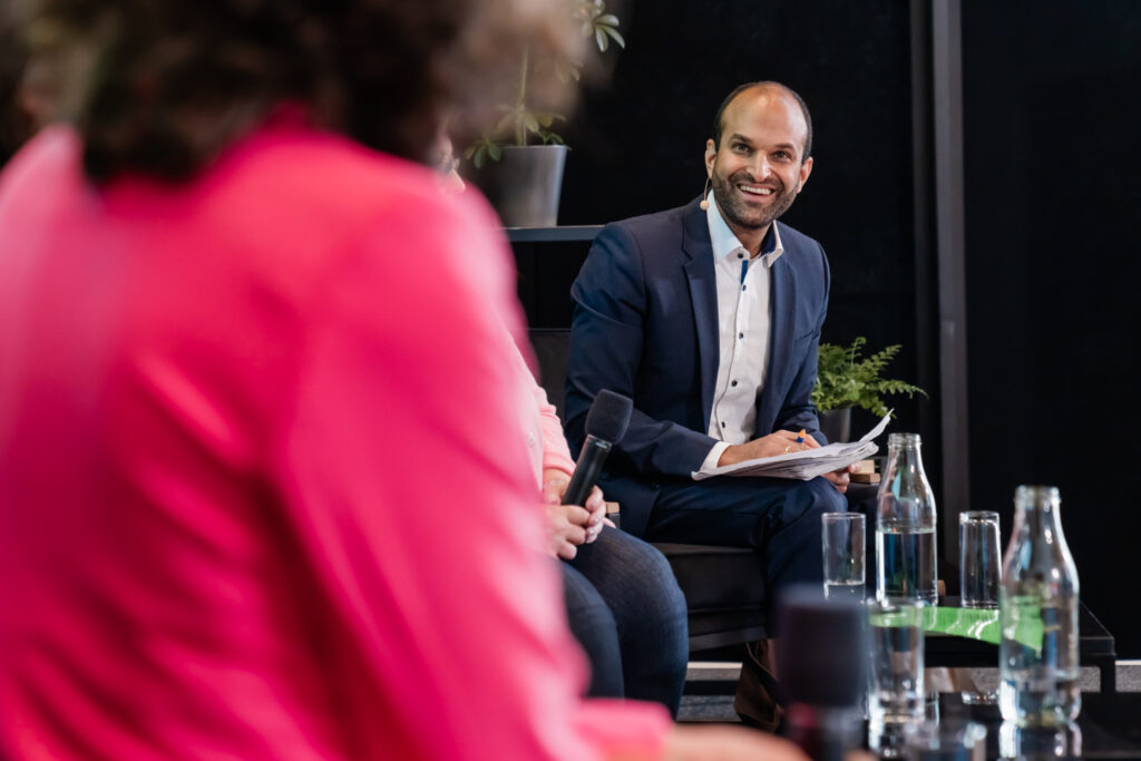Das Bild zeigt eine Podiumsdiskussion oder ein Panel-Gespräch. Im Fokus steht ein Mann in einem dunkelblauen Anzug mit einem weißen Hemd, der ein Headset-Mikrofon trägt. Er hält Notizen in der Hand und lächelt, während er aufmerksam der Diskussion folgt. Vor ihm stehen mehrere Wasserflaschen und Gläser auf einem Tisch. Im Vordergrund sind unscharf zwei weitere Personen zu sehen, eine in einem pinken Blazer, die gerade spricht, und eine weitere Person mit einem Handmikrofon. Die Szenerie wirkt professionell und freundlich, mit Pflanzen im Hintergrund, die eine angenehme Atmosphäre schaffen.