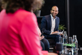 Das Bild zeigt eine Podiumsdiskussion oder ein Panel-Gespräch. Im Fokus steht ein Mann in einem dunkelblauen Anzug mit einem weißen Hemd, der ein Headset-Mikrofon trägt. Er hält Notizen in der Hand und lächelt, während er aufmerksam der Diskussion folgt. Vor ihm stehen mehrere Wasserflaschen und Gläser auf einem Tisch. Im Vordergrund sind unscharf zwei weitere Personen zu sehen, eine in einem pinken Blazer, die gerade spricht, und eine weitere Person mit einem Handmikrofon. Die Szenerie wirkt professionell und freundlich, mit Pflanzen im Hintergrund, die eine angenehme Atmosphäre schaffen.