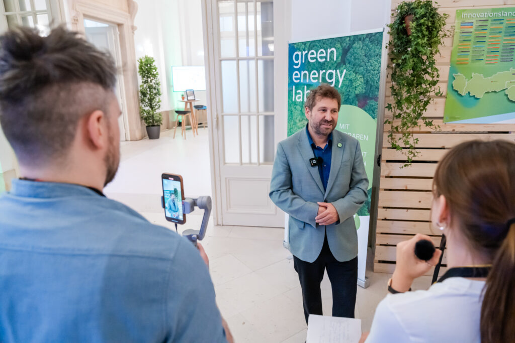 Das Bild zeigt ein Interview, das bei einer Veranstaltung im Green Energy Lab geführt wird. Ein Mann in einem hellblauen Anzug mit einem Mikrofonclip steht vor einem grünen Banner mit dem Schriftzug 