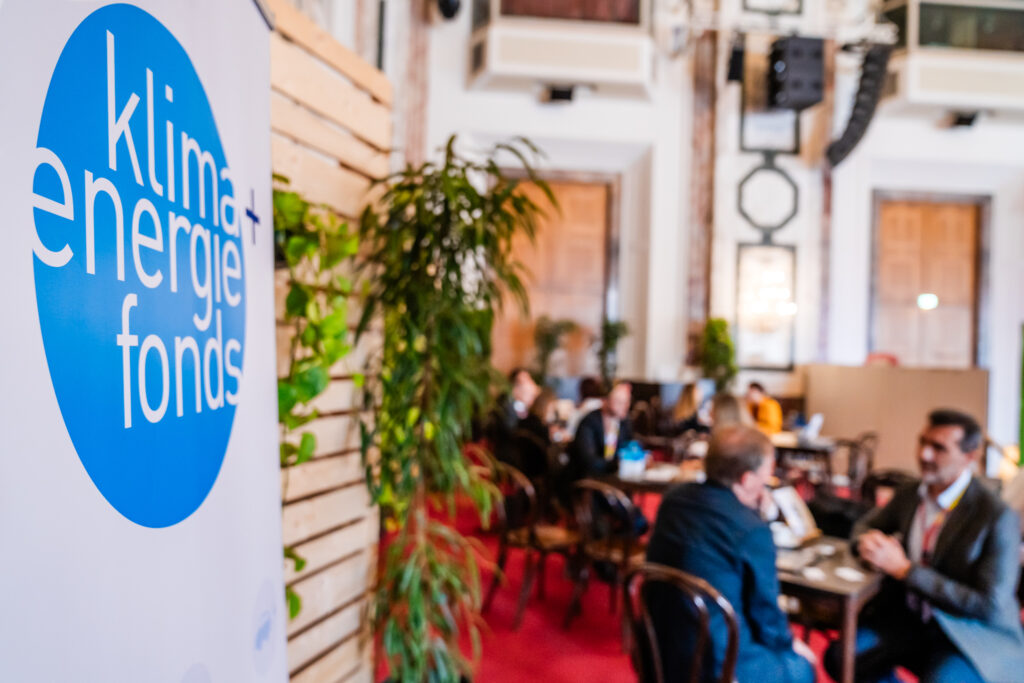 Ein Banner mit dem Logo des 'Klima + Energie Fonds' ist im Vordergrund zu sehen. Dahinter befinden sich mehrere Personen in einem Networking-Bereich, die in Gespräche an kleinen Tischen vertieft sind. Die Umgebung ist mit Pflanzen dekoriert und der Raum hat eine elegante Einrichtung mit hohen Decken, Holzvertäfelungen und einem roten Teppichboden.