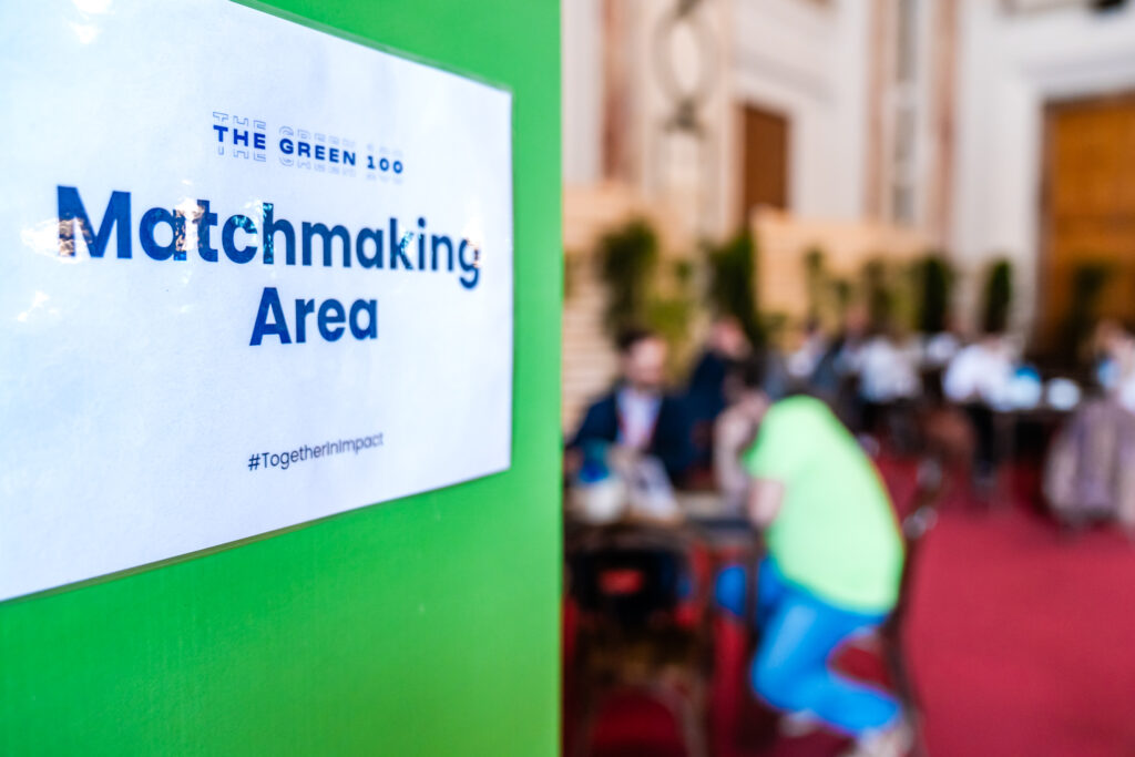 Ein Schild mit der Aufschrift 'THE GREEN 100 - Matchmaking Area #TogetherInImpact' ist im Vordergrund an einer grünen Wand angebracht. Im unscharfen Hintergrund sitzen mehrere Personen in einer offenen Networking-Umgebung an Tischen, vertieft in Gespräche. Die Atmosphäre wirkt geschäftig und professionell, mit einer roten Teppichfläche und Pflanzen, die den Raum dekorieren.