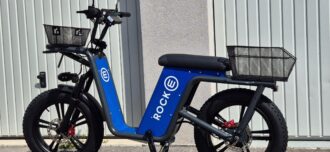 Ein blaues einspuriges Elektrofahrzeug mit großen Reifen und einem Korb vorne und hinten. Das Fahrzeug steht vor einem grauen Garagentor.