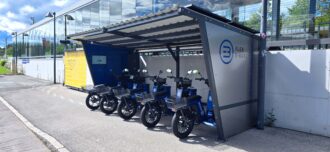 Reihe blauer einspuriger E-Fahrzeuge mit Körben, geparkt unter einem Dach neben einem digitalen Bildschirm. Reihe blauer einspuriger E-Fahrzeuge mit Körben, geparkt unter einem Dach neben einem digitalen Bildschirm.