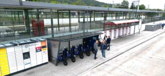 Vor einem Bahnhof wird eine Sharing-Station für einspurige E-Fahrzeuge gezeigt, die zwischen einer Bushaltestelle und einer Paketstation befindet. Davor stehen Menschen, die sich unterhalten.