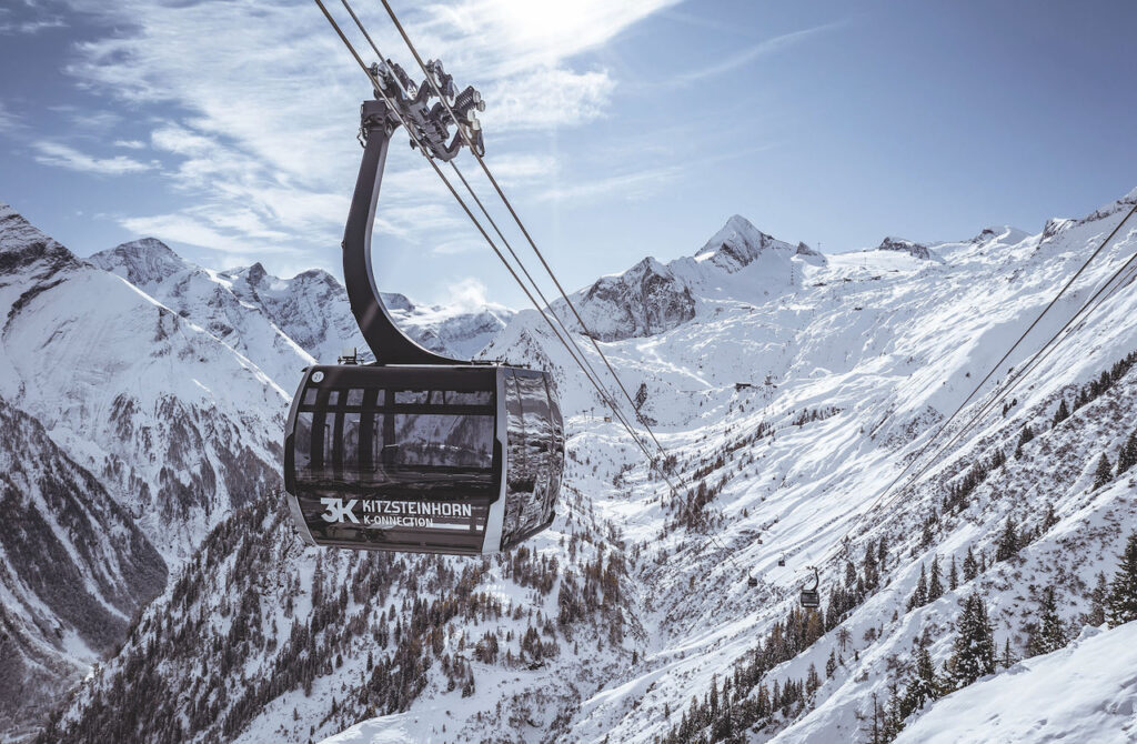 Eine moderne Gondel der 3K K-onnection schwebt über eine verschneite Alpenlandschaft am Kitzsteinhorn. Im Hintergrund erstreckt sich eine atemberaubende Bergkulisse mit schneebedeckten Gipfeln unter einem klaren blauen Himmel. Die Seilbahn verläuft über dichte Nadelwälder, die teils von Schnee bedeckt sind, und bietet einen spektakulären Blick auf das alpine Winterparadies.