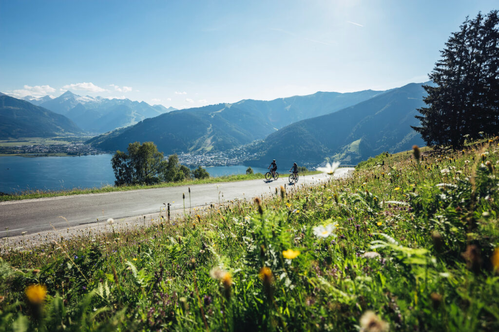 Zwei Radfahrer fahren auf einer kurvigen Bergstraße mit Blick auf einen malerischen See und umliegende grüne Berge. Im Vordergrund blühen bunte Wildblumen, während im Hintergrund ein idyllisches Tal mit einem kleinen Ort am Wasser zu sehen ist. Der Himmel ist klar und sonnig, was die sommerliche, frische Atmosphäre der alpinen Landschaft unterstreicht.