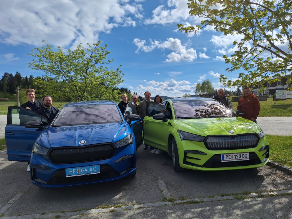 Zwei bunte E-Autos, die nebeneinander unter einem sonnigen Himmel geparkt sind und neben welchen ein paar Leute stehen.