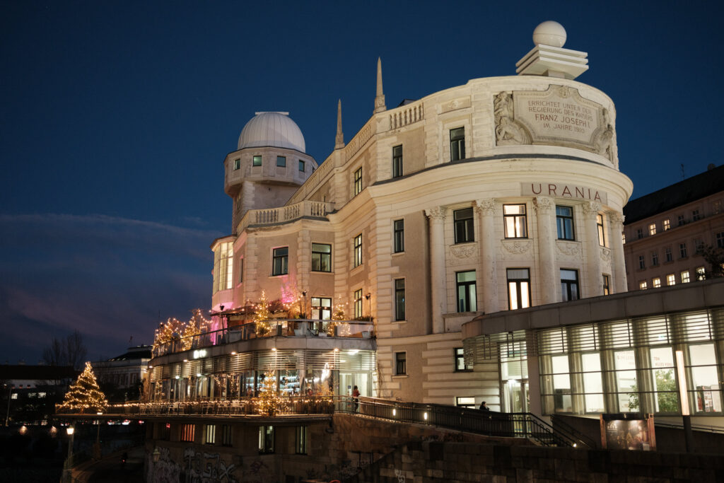 Das Gebäude der Wiener Urania bei Nacht, festlich beleuchtet mit Lichterketten und dekorierten Weihnachtsbäumen auf der Terrasse. Die historische Fassade mit der Aufschrift „URANIA“ und der Inschrift „ERRICHTET UNTER DER REGIERUNG DES KAISERS FRANZ JOSEPH I. IM JAHRE 1910“ ist gut sichtbar. Im Vordergrund führt eine beleuchtete Rampe zum Eingang, während der Himmel im Hintergrund dunkelblau ist. Die Architektur mit dem markanten Kuppelturm und den verzierten Fassadenelementen steht im Kontrast zur modernen Glasfassade im unteren Bereich.