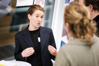 Eine Frau im dunklen Blazer spricht mit zwei anderen Personen, die mit dem Rücken zur Kamera stehen. Sie gestikuliert mit den Händen, während sie spricht, und wirkt engagiert und konzentriert. Das Gespräch findet an einem Stehtisch in einem hellen Raum statt, vermutlich im Rahmen einer Networking-Veranstaltung. Im Hintergrund sind Poster und Fenster zu sehen.
