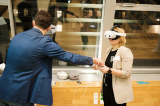 Eine Frau trägt eine Virtual-Reality-Brille und hält einen Controller in der Hand, während sie mit einem Mann in einem blauen Anzug interagiert. Der Mann, der mit dem Rücken zur Kamera steht, streckt seine Hand aus, als würde er der Frau etwas überreichen oder eine Anweisung geben. Im Hintergrund spiegeln sich andere Personen in den großen Fenstern. Auf einer Holzablage liegen VR-Zubehör, ein Glas Wasser und einige Prospekte.