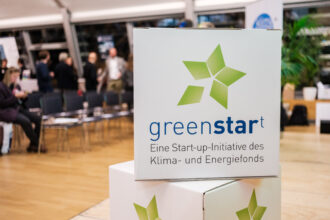 Ein weißer Würfel mit dem Logo und der Aufschrift „greenstart – Eine Start-up-Initiative des Klima- und Energiefonds“ steht im Vordergrund. Das grüne Sternsymbol ist prominent platziert. Im Hintergrund sind Stuhlreihen und mehrere Menschen zu sehen, die sich unterhalten, was auf eine Veranstaltung oder Networking-Situation hindeutet. Rechts im Bild sind Pflanzen als Teil der Dekoration erkennbar.