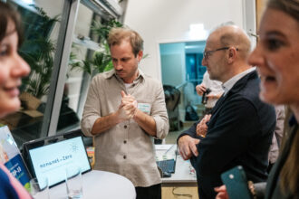Ein Mann mit einem Namensschild auf seinem Hemd steht an einem Tisch und erklärt etwas, während er die Hände ineinander verschränkt. Vor ihm ein Laptop mit dem Schriftzug „nonanet_zero“ und dem Slogan „we bring the workspace for climate goals“. Zwei Zuhörer, einer mit Brille, hören aufmerksam zu, während im Vordergrund eine Frau unscharf ins Bild ragt. Im Hintergrund hält eine Person eine Kamera.