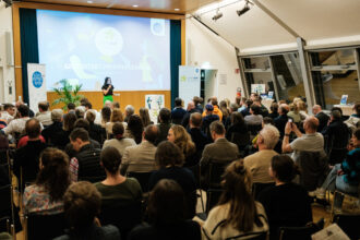 Ein Veranstaltungsraum voller Menschen, die aufmerksam einer Frau auf der Bühne zuhören. Sie trägt ein Mikrofon und spricht vor einer großen Leinwand mit dem Schriftzug „GREENSTART PREISVERLEIHUNG – Wer sind die TOP-37?“. Links und rechts stehen Banner mit dem Logo des Klima- und Energiefonds. Einige Personen im Publikum machen Fotos oder nehmen die Veranstaltung mit dem Smartphone auf.