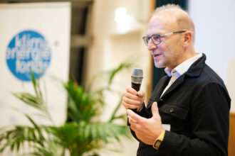 Ein Mann mit Brille und einem schwarzen Jackett spricht in ein Mikrofon bei einer Veranstaltung. Hinter ihm steht ein Banner mit dem Logo des „Klima- und Energiefonds“. Eine grüne Pflanze ist im Vordergrund zu sehen.