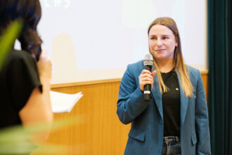 Eine junge Frau mit langen, glatten Haaren spricht bei einer Veranstaltung ins Mikrofon. Sie trägt einen blauen Blazer über einem schwarzen Shirt und Jeans. Im Vordergrund ist eine andere Person mit dunkler Kleidung zu sehen, die ein Mikrofon hält und ein Notizbuch in der Hand hat. Der Hintergrund zeigt eine Präsentationsleinwand mit leicht verschwommenem Text und Grafiken.