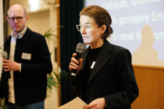 Eine Frau im dunklen Blazer spricht auf einer Bühne in ein Mikrofon und hält ein Blatt Papier in der Hand. Ihr Gesicht wird teilweise von der Projektion einer Leinwand beleuchtet, auf der Text zu sehen ist. Im Hintergrund steht ein Mann mit Brille und Mikrofon, der aufmerksam zuhört. Links im Bild sind große Pflanzen zu sehen, die zur Bühnendekoration gehören. Die Szene wirkt wie ein Präsentations- oder Pitch-Moment im Rahmen einer Veranstaltung.