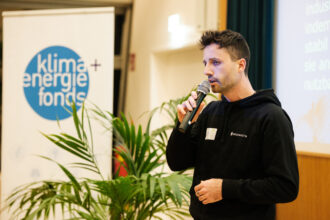 Ein Mann im schwarzen Hoodie mit dem Logo „sequestra“ spricht in ein Mikrofon. Er steht auf einer Bühne, umgeben von Pflanzen. Im Hintergrund befindet sich ein Roll-up-Banner mit dem Logo des „klima + energie fonds“ und ein Teil einer Leinwand, auf der Text und grafische Elemente zu sehen sind. Der Mann wirkt konzentriert, während er zu einem Publikum spricht.
