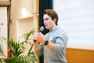 Ein junger Mann mit braunen Haaren und einem hellblauen Pullover steht auf einer Bühne und spricht in ein Mikrofon. Er trägt eine Smartwatch und ein Namensschild, während er mit einer Hand gestikuliert. Im Hintergrund ist eine Leinwand mit einer Präsentation zu sehen, auf der das Wort „Lösung“ lesbar ist. Daneben befinden sich eine grüne Pflanze und ein Banner mit dem „Klima + Energie Fonds“-Logo.
