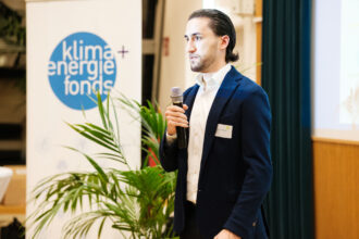 Ein junger Mann mit zurückgekämmten Haaren und einem blauen Blazer steht auf einer Bühne und spricht in ein Mikrofon. Er trägt ein Namensschild und schaut konzentriert in Richtung Publikum. Im Hintergrund ist ein Banner mit dem Logo des „Klima + Energie Fonds“ sowie eine Projektionsfläche und eine grüne Pflanze zu sehen.