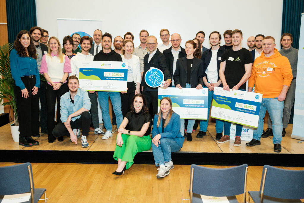 Eine große Gruppe von Menschen steht gemeinsam auf einer Bühne und lächelt in die Kamera. Drei Personen in der ersten Reihe halten übergroße Schecks mit den Aufschriften „sequestra“, „Zukunft Bestand“ und „TerraBreeze by Social Cooling“, jeweils mit dem Betrag „EUR 20.000,-“ und dem Titel „TOP-3 GREENSTART 2024“. Zwei Frauen sitzen vorne auf der Bühnenkante, während der Rest der Gruppe dahinter steht. Links und rechts im Hintergrund befinden sich Roll-up-Banner der „greenstart“-Initiative des Klima- und Energiefonds Österreichs. Die Gruppe besteht aus den Gewinnerinnen, Organisatorinnen und weiteren Beteiligten der Preisverleihung.
