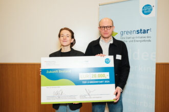 Eine Frau und ein Mann stehen nebeneinander und halten gemeinsam einen übergroßen Scheck mit der Aufschrift „Zukunft Bestand“ und dem Betrag „EUR 20.000,-“ für die Auszeichnung als „TOP-3 GREENSTART 2024“. Im Hintergrund befindet sich ein Roll-up-Banner der „greenstart“-Initiative des Klima- und Energiefonds Österreichs mit dem grünen Stern-Logo und Schlagwörtern wie „Idee“, „Networking“ und „Erneuerbar“. Beide blicken lächelnd in die Kamera.