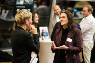 Zwei Frauen führen ein Gespräch bei einer Networking-Veranstaltung. Die Frau auf der rechten Seite trägt eine Brille, ein bordeauxfarbenes Blazer und eine schwarze Bluse, während sie mit einer lebhaften Gestik spricht. Die andere Frau, mit kurzem blondem Haar und großen goldenen Ohrringen, hört aufmerksam zu und hält ein paar Dokumente in der Hand. Im Hintergrund unterhalten sich weitere Gäste an Stehtischen mit Getränken.
