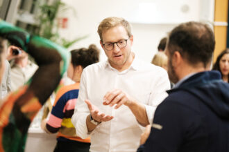 Ein Mann mit Brille und weißem Hemd spricht gestikulierend mit einer anderen Person, die eine dunkelblaue Jacke trägt. Die Unterhaltung findet in einer lebhaften Veranstaltung statt, im Hintergrund sind weitere Teilnehmende, darunter eine Person mit einem bunten gestreiften Pullover, sichtbar. Die Atmosphäre wirkt kommunikativ und engagiert.