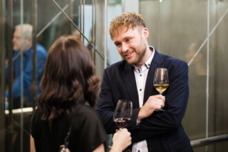 Ein Mann mit lockigem, blondem Haar und einem dunklen Sakko hält ein Glas Weißwein und lächelt freundlich, während er sich mit einer Frau unterhält. Die Frau, deren Gesicht nicht sichtbar ist, hat braunes, welliges Haar und trägt ein schwarzes Outfit, während sie ein Glas Rotwein hält. Die Unterhaltung findet in einer stilvollen, modernen Umgebung mit Glaswänden und Spiegelungen statt, die eine gesellige Atmosphäre vermitteln.