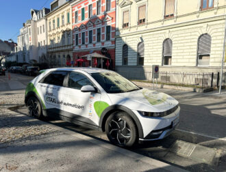 Ein weißes Elektro-Taxi mit grünen Akzenten, das auf einer Stadtstraße geparkt ist, auf dem steht: „Dieses eTAXI lädt automatisiert!“