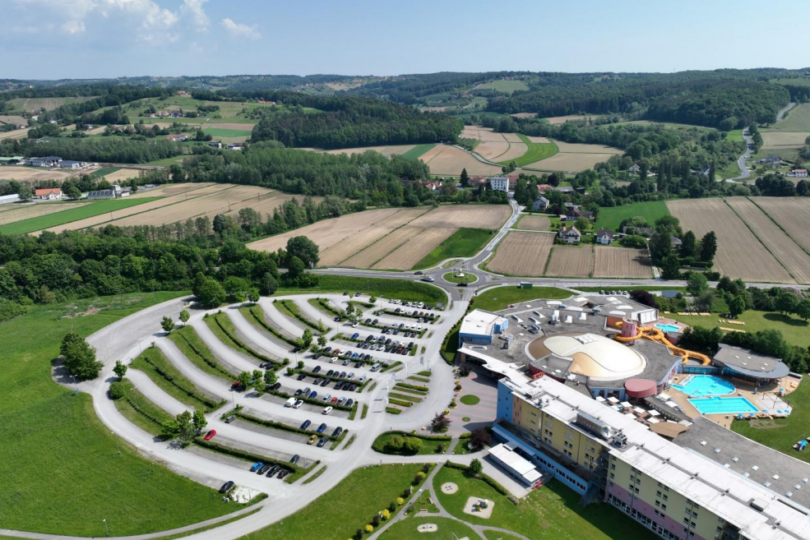 Luftaufnahme eines geschwungenen Gebäudekomplexes mit angrenzendem Parkplatz und Schwimmbad, umgeben von Feldern.