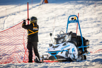 Person in Skiausrüstung, die ein rotes Sicherheitsnetz in der Nähe eines Schneemobils auf einer verschneiten Piste aufspannt.