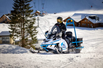 Eine Person, die an einem sonnigen Tag in der Nähe von Skiliften und Berghütten mit einem Motorschlitten fährt.