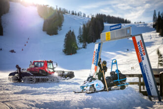 Ein Pistenfahrzeug und ein Motorschlitten, die in der Nähe eines Skiliftschilds an einem verschneiten Hang unter klarem Himmel geparkt sind.