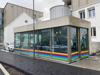 Ein modernes Schaufenster mit großen Fenstern, die unten mit einem regenbogenfarbenen Aufkleber und der Aufschrift „MOBILITY“ versehen sind.