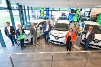 Eine Gruppe von Menschen steht neben Elektrofahrzeugen an einer Ladestation in einem modernen Gebäude.