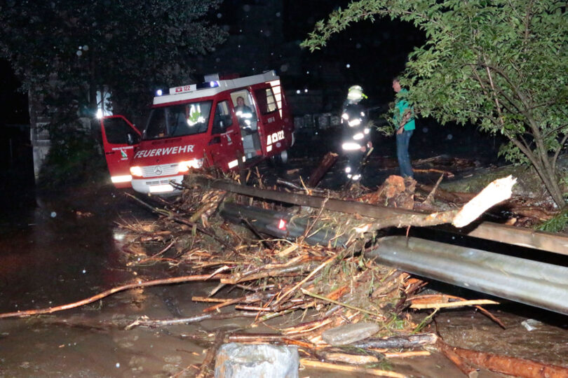 Feuerwehreinsatz bei Nacht nach starkem Unwetter: Ein Feuerwehrfahrzeug steht auf einer überfluteten Straße voller Treibholz und Äste. Einsatzkräfte in Schutzkleidung sichern die Umgebung und beseitigen die Schäden.