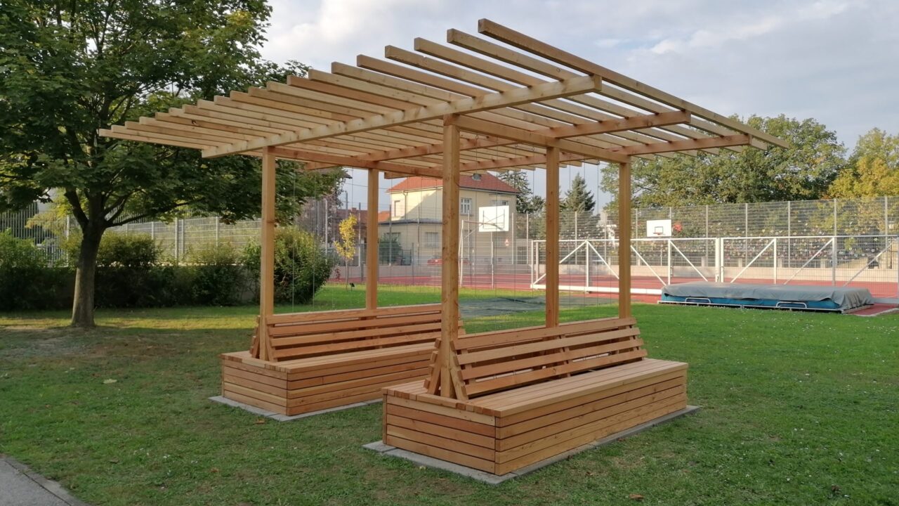 Die Aufnahme zeigt eine neu errichtete Holzpergola mit integrierten Sitzbänken auf einer Rasenfläche im Außenbereich einer Schule. Die Pergola besteht aus hellem, unbehandeltem Holz und bietet Sitzgelegenheiten auf allen vier Seiten. Im Hintergrund ist ein umzäunter Sportplatz mit Basketballkörben und einer Hochsprunganlage zu sehen. Links steht ein großer Laubbaum, rechts und im Hintergrund weitere Bäume und Gebäude. Die Szene wirkt ruhig und gepflegt – vermutlich handelt es sich um eine begrünbare Pergola oder ein Outdoor-Klassenzimmer im Rahmen eines Schulprojekts.