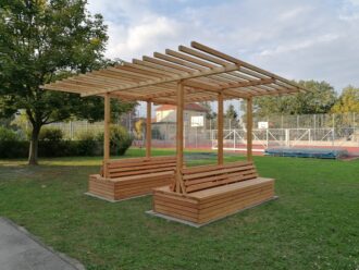 Die Aufnahme zeigt eine neu errichtete Holzpergola mit integrierten Sitzbänken auf einer Rasenfläche im Außenbereich einer Schule. Die Pergola besteht aus hellem, unbehandeltem Holz und bietet Sitzgelegenheiten auf allen vier Seiten. Im Hintergrund ist ein umzäunter Sportplatz mit Basketballkörben und einer Hochsprunganlage zu sehen. Links steht ein großer Laubbaum, rechts und im Hintergrund weitere Bäume und Gebäude. Die Szene wirkt ruhig und gepflegt – vermutlich handelt es sich um eine begrünbare Pergola oder ein Outdoor-Klassenzimmer im Rahmen eines Schulprojekts.